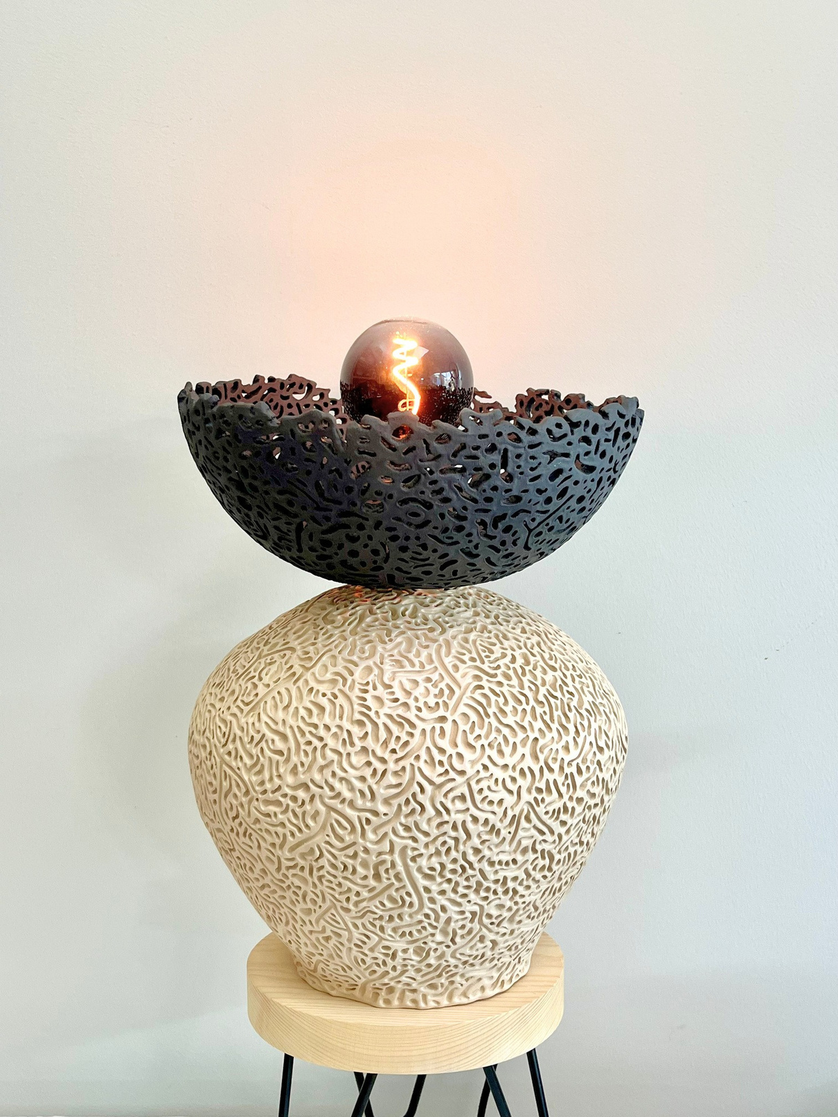 Nyx halo lamp sculpture, dark stoneware perforated bowl balanced on a pale textured sphere, with glowing filament bulb — view 3