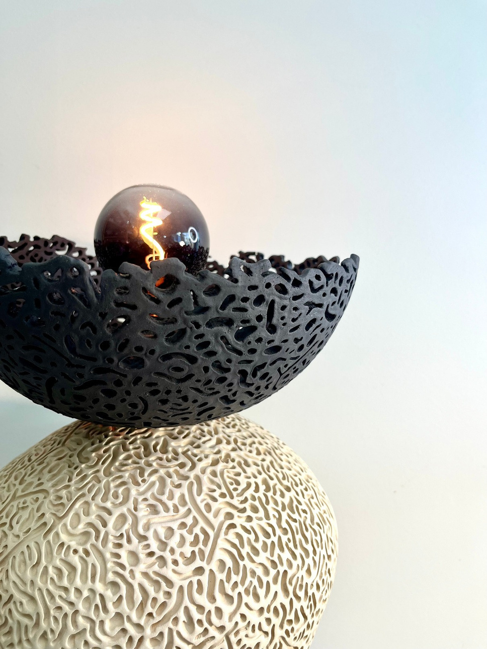 Nyx halo lamp sculpture, dark stoneware perforated bowl balanced on a pale textured sphere, with glowing filament bulb